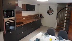 Una cocina moderna con electrodomésticos integrados, una zona de comedor con una mesa lista para dos personas, y una escalera que conduce a un nivel superior.