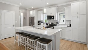 A modern kitchen with white cabinetry, a central island with stools, stainless steel appliances, and pendant lighting. - Sandy Feet Retreat at Lost Key Villas (Pensacola)