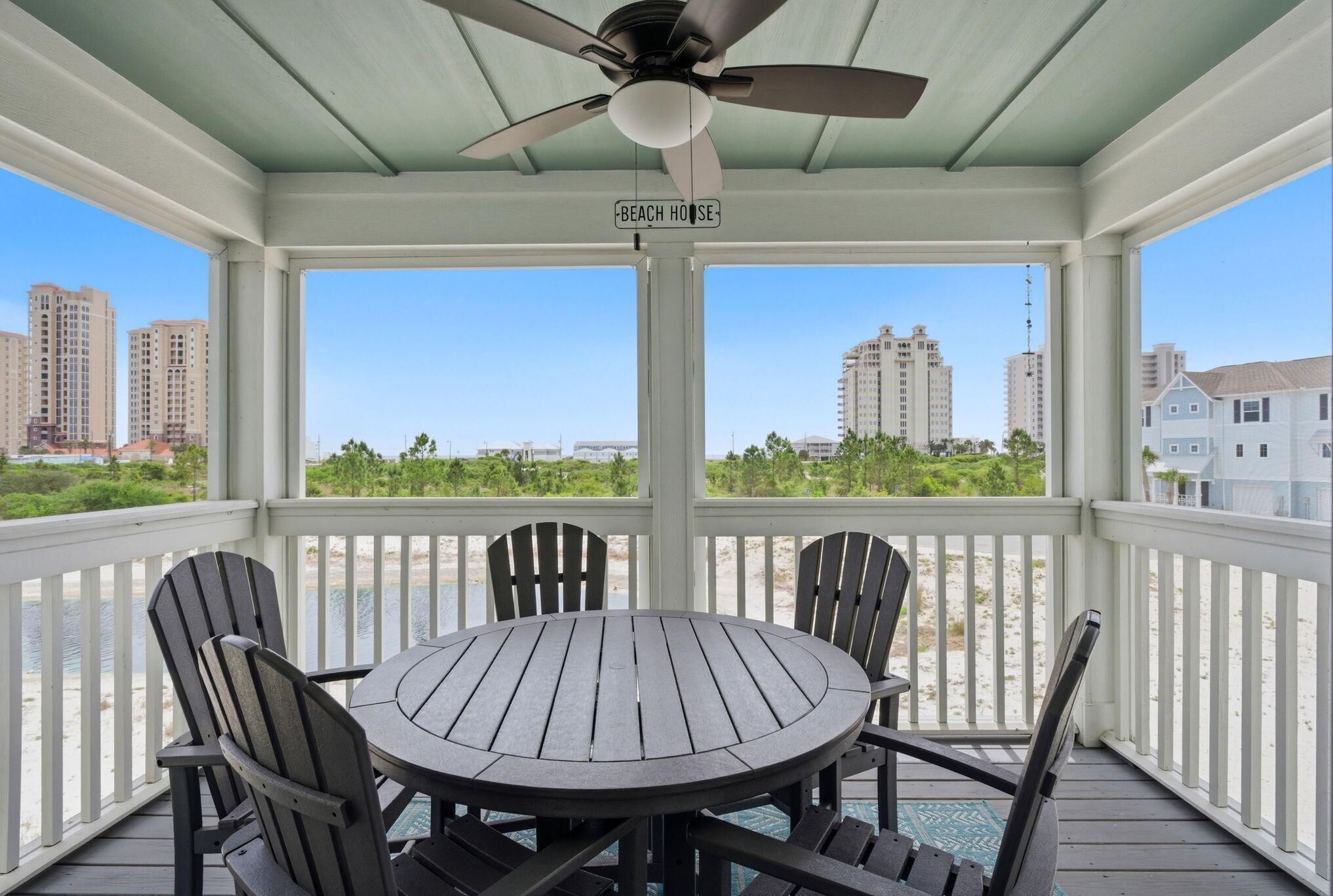 A covered outdoor seating area with a table and chairs, a ceiling fan, and a view of the beach and buildings.