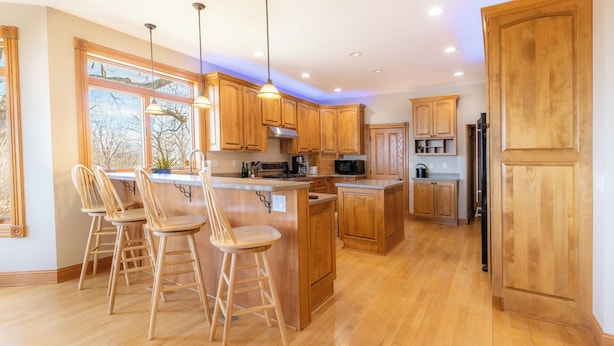 Una cocina moderna con armarios de madera, una isla central con taburetes, iluminación colgante y vistas amplias a través de grandes ventanales.
