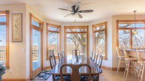 Un comedor con una amplia mesa de madera, sillas, un ventilador de techo y ventanas grandes con vistas a árboles.