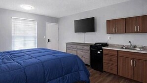 A bedroom with a bed, a TV, a kitchen area with cabinets, and a window with blinds.