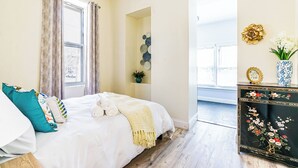 Een slaapkamer met een bed, kussens, een nachtkastje met een vaas en een raam met gordijnen.