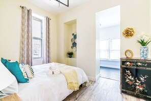 Ein Schlafzimmer mit einem Bett, Kissen, einem Nachttisch mit einer Vase und einem Fenster mit Vorhängen.