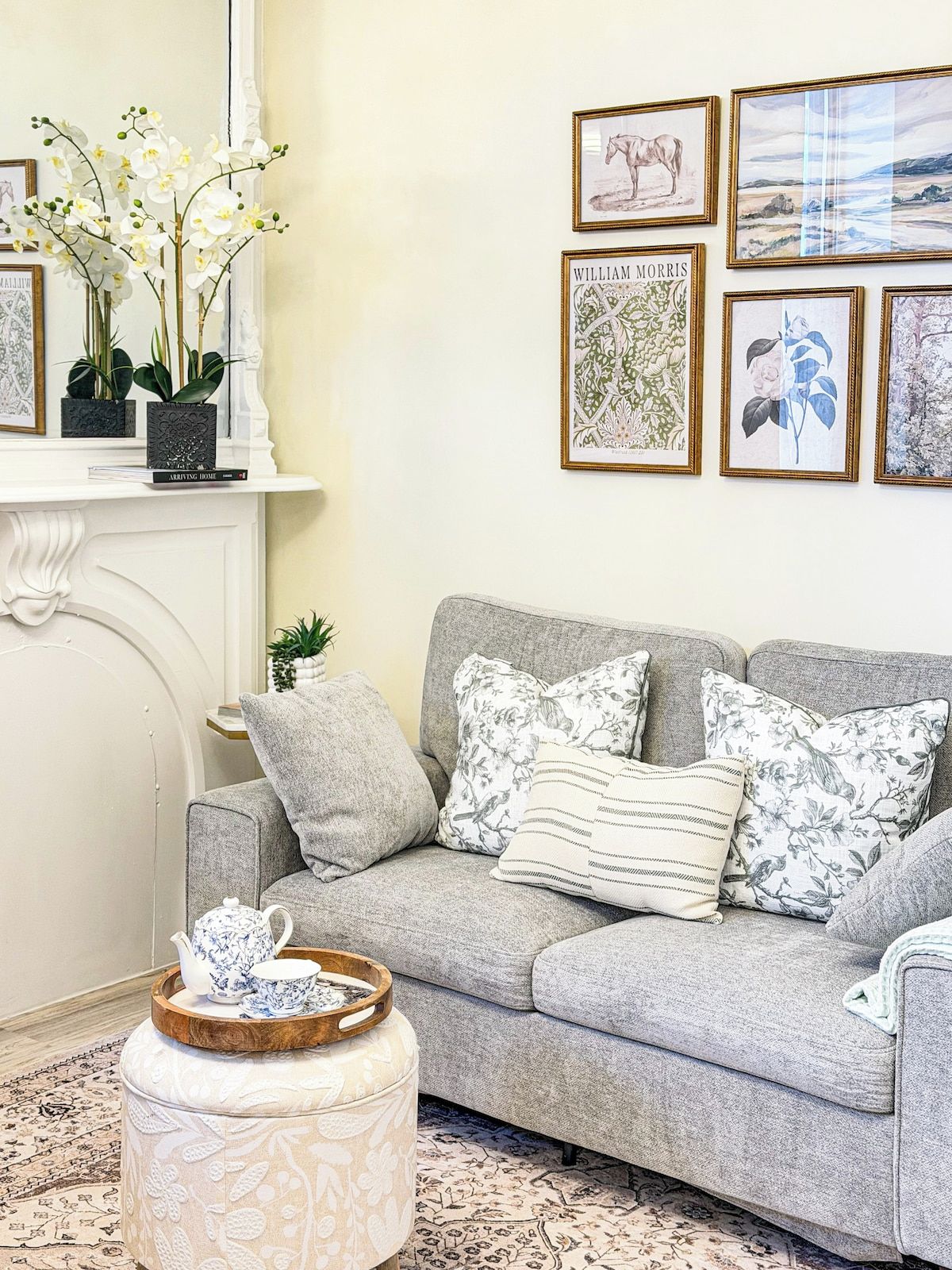 A living room with a grey sofa, floral cushions, a small round ottoman, and a fireplace with a white mantel.