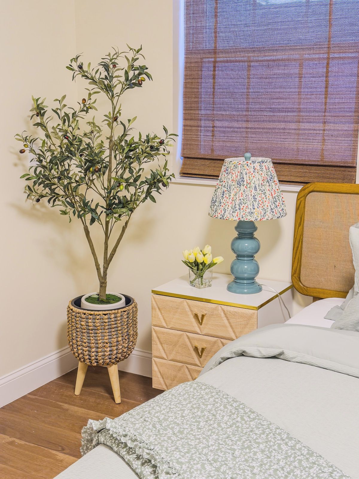 A bedroom with a bed, a nightstand with a lamp, a small potted plant, and a window with blinds.