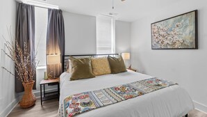 Une chambre à coucher comprenant un lit, des lampes de chevet, un ventilateur de plafond, une fenêtre avec des rideaux, un tableau accroché au mur et un vase décoratif.
