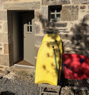 A yellow backpack and a red item on a wooden bench outside a stone building with a door and a small window.