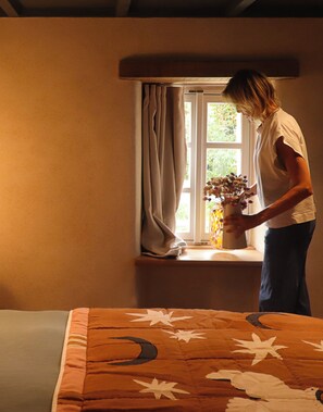 Una persona de pie junto a una ventana con cortinas, sosteniendo un florero con flores.