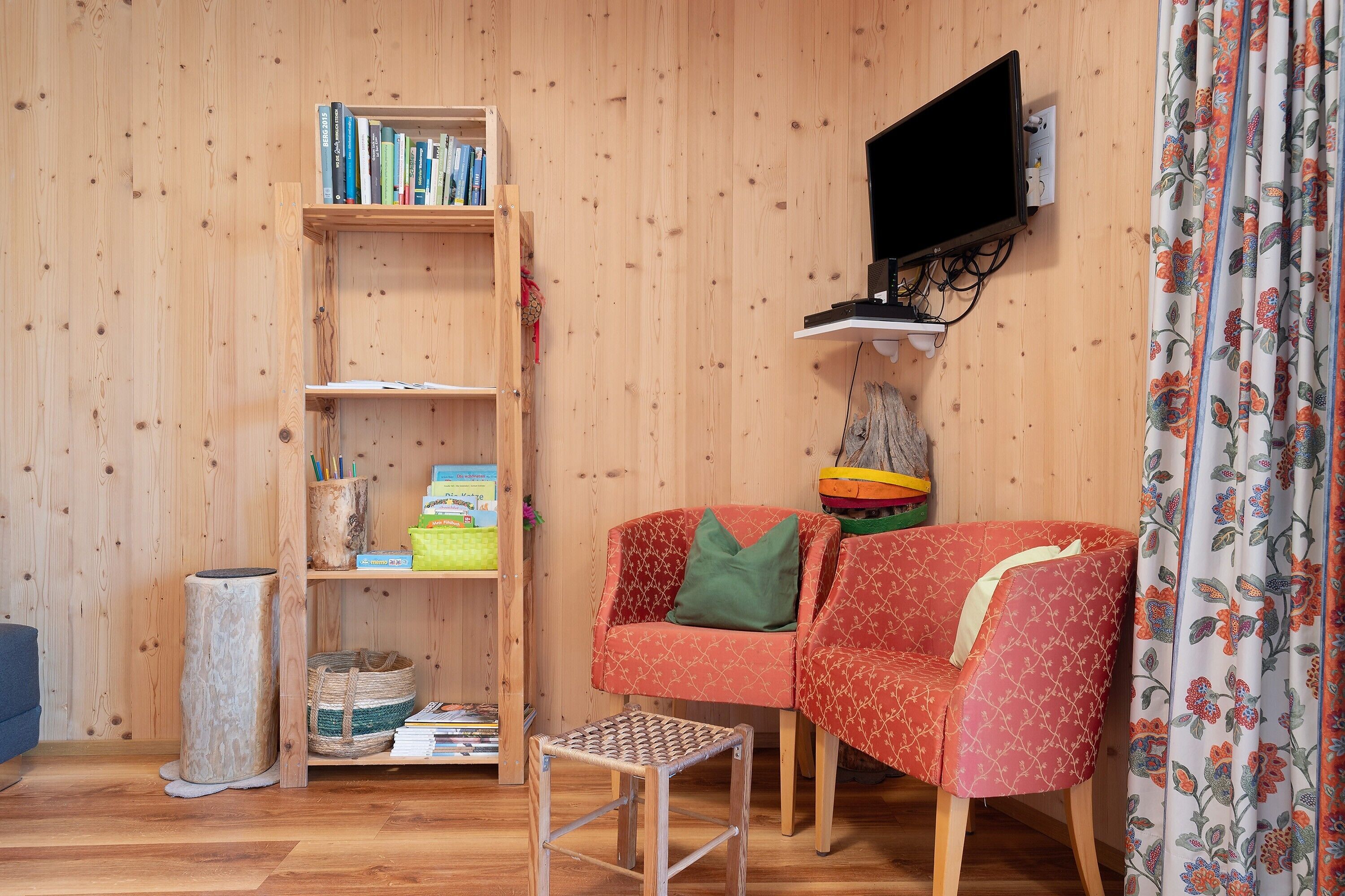 Une pièce comprenant une bibliothèque, un téléviseur, un canapé rouge à motifs, un petit tabouret en bois et un rideau orné de motifs floraux.