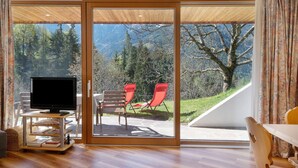 A room with a television on a stand, two red chairs, and a sliding glass door leading to an outdoor area with a view of trees and mountains. - Chalet 'Petrunderhof Herbstparadies' with Mountain View, Shared Terrace and Wi-Fi (Villnöß)