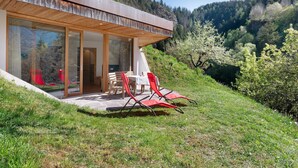 A modern house with a glass facade, a wooden deck, and a dining area with red chairs. - Chalet 'Petrunderhof Herbstparadies' with Mountain View, Shared Terrace and Wi-Fi (Villnöß)