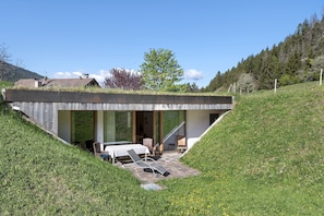 Una casa moderna con techo inclinado, un patio con mobiliario exterior y vistas del paisaje circundante.