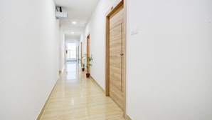 A long, narrow hallway with a polished floor, wooden doors, and potted plants. - Saver Single Karmanghat Midtown (Hyderabad)