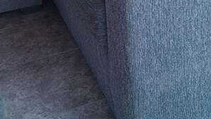 Un sofá seccional gris con un cojín de color oscuro, una cortina verde y un suelo de baldosas.