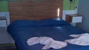 Una cama con colcha azul y toallas blancas, cabecera de madera, mesita de noche con lámpara y un enchufe de pared.