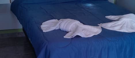 A bed with a blue bedspread and white towels, a wooden headboard, a nightstand with a lamp, and a wall socket.