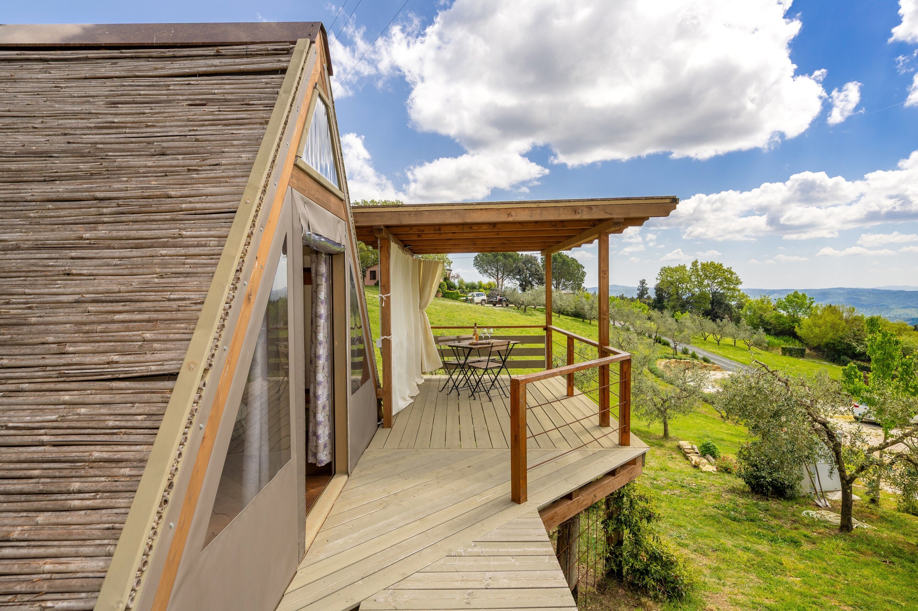 Un terrazzo in legno con tavolo e sedie, affacciato su un paesaggio verdeggiante con alberi e una vista lontana.
