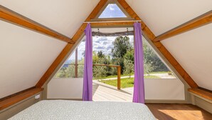 A triangular attic bedroom with a wooden ceiling, a bed, and a view of a garden.