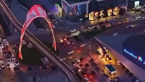Um hotel com vários andares e um destaque para o letreiro 'Blue Sky', rodeado por uma movimentada rua urbana com uma ponte de arco vermelha marcante.