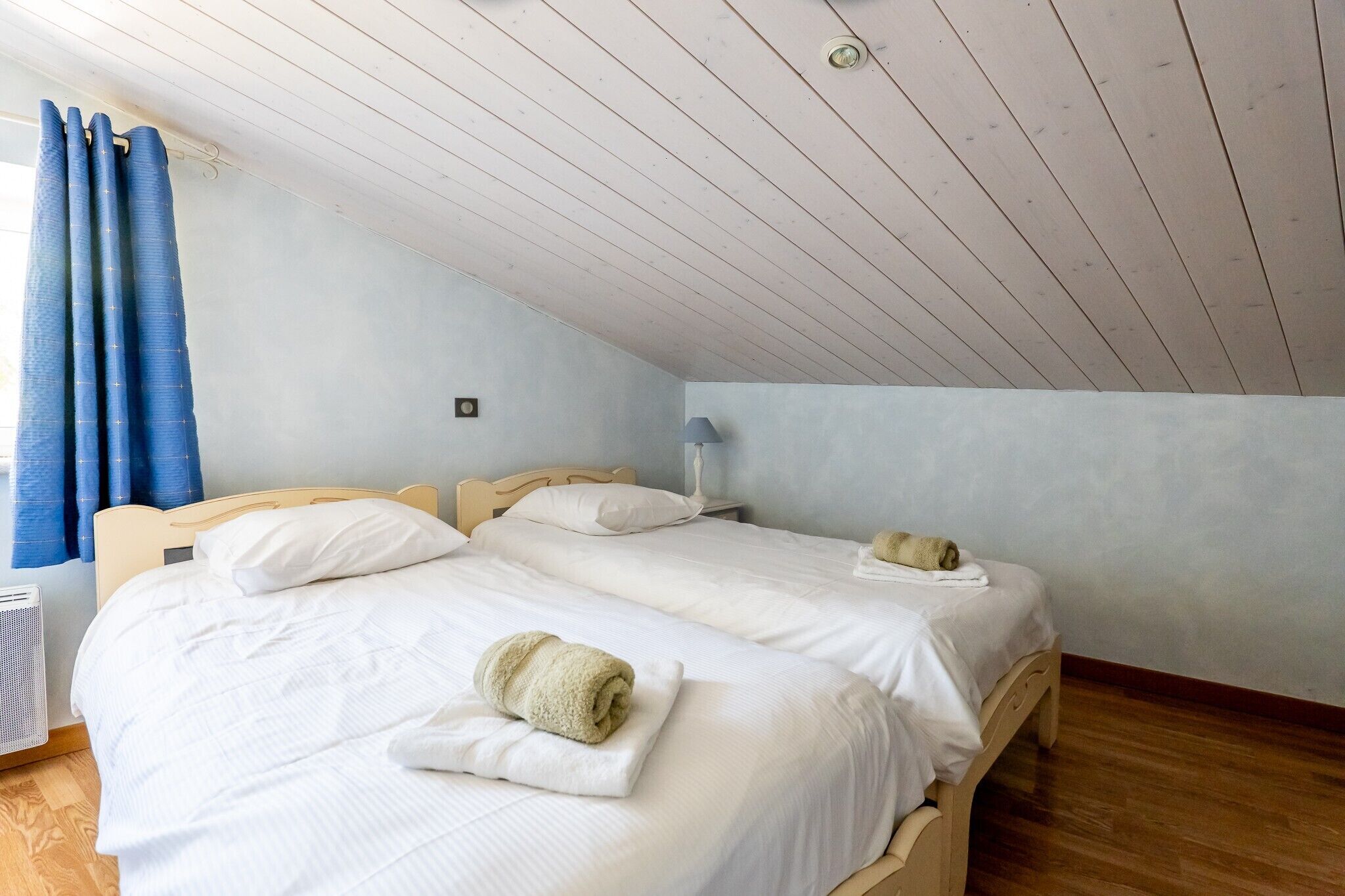 Una habitación con dos camas, una ventana con cortinas azules, una lámpara de pared y un radiador.
