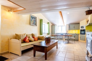 A cozy living room with a sofa, coffee table, and dining area with chairs. A kitchen with cabinets and appliances is visible in the background. - Le Mas des Oliviers – Peaceful 2-Level Apartment with Pool, Nature Views, and Shared Amenities (Redortiers)