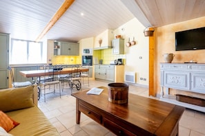 A spacious kitchen with a dining area, wooden ceiling beams, a television, and a fireplace. - Le Mas des Oliviers – Peaceful 2-Level Apartment with Pool, Nature Views, and Shared Amenities (Redortiers)