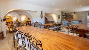 Un comedor amplio con una larga mesa de madera y sillas metálicas, rodeado de arquerías y una zona de bar con estantes.