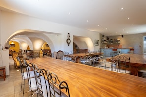 A spacious dining area with a long wooden table and metal chairs, a bar area with shelves, and a clock on the wall.