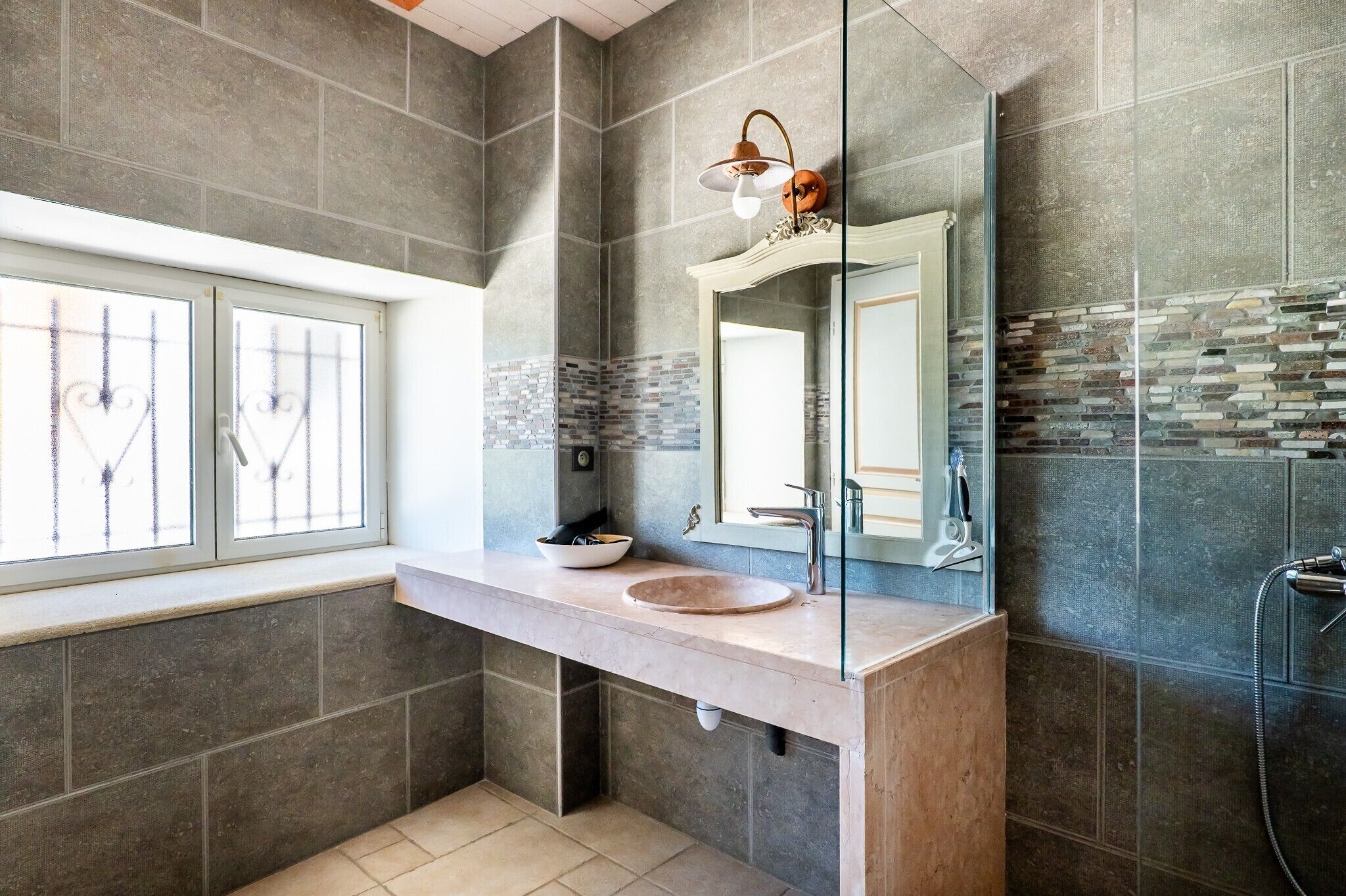 Un baño con un espejo grande, un lavamanos, una ducha y una ventana con barrotes.