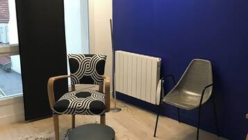 A room with a blue wall, a radiator, a window with curtains, a chair, a table, and a patterned rug.