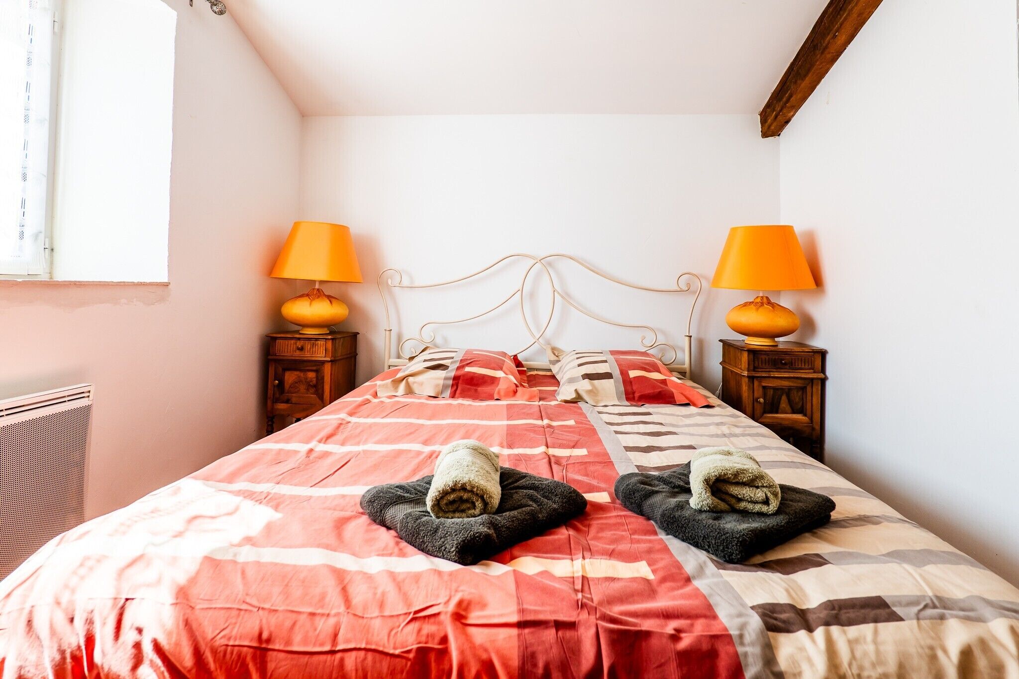 Una cama con colcha roja y beige, dos lámparas en mesitas de noche de madera, y una ventana con cortinas.