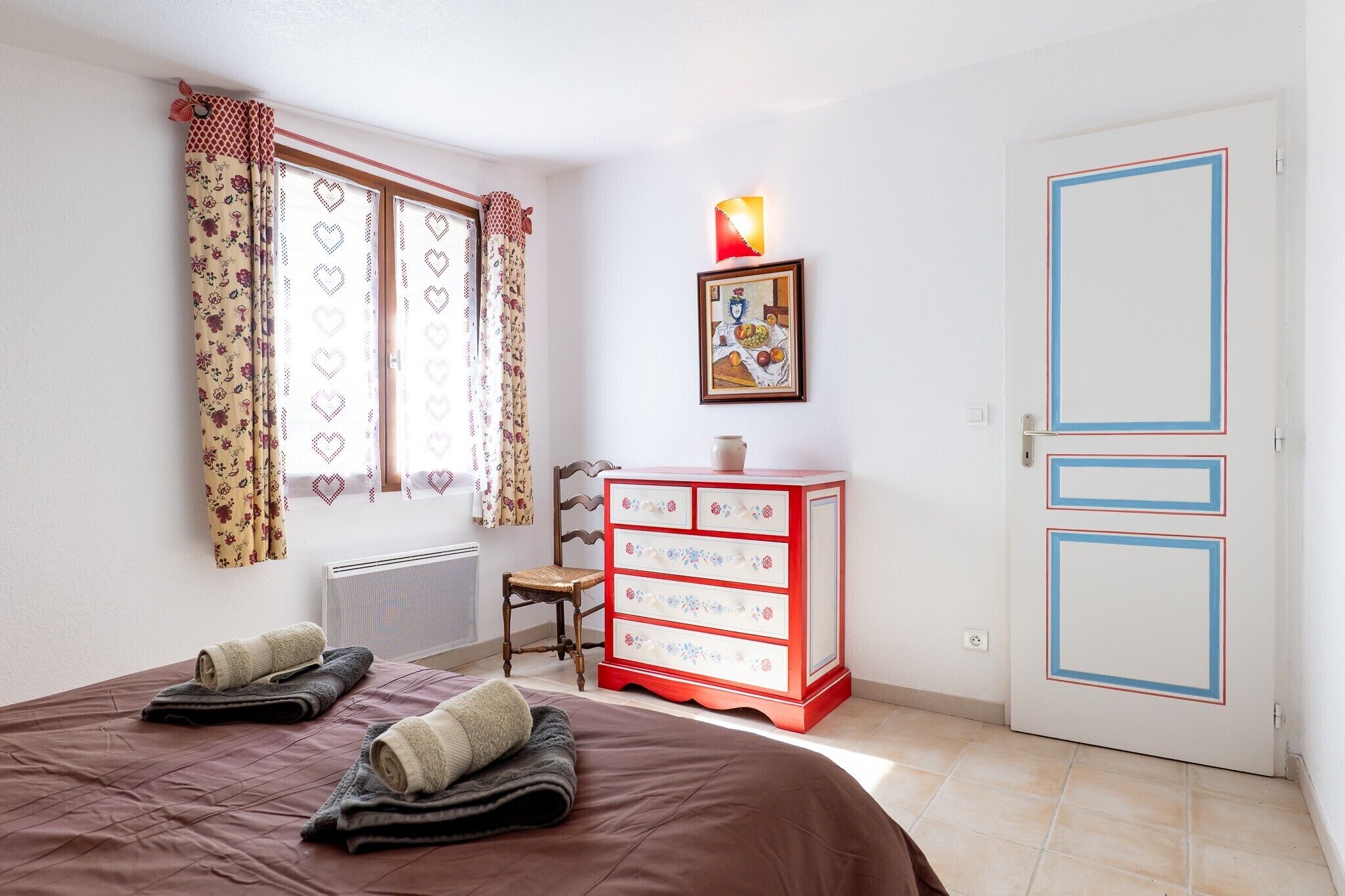 Camera da letto con un letto, una cassettiera, una sedia, un termosifone, una finestra con tende e una porta.