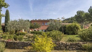 A large house with a stone wall in the foreground, surrounded by various plants and trees. - Magnificent Provençal villa in a quiet private domain (Tourtour)