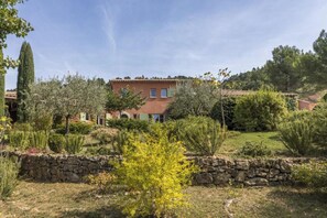 A large house with a stone wall in the foreground, surrounded by various plants and trees. - Magnificent Provençal villa in a quiet private domain (Tourtour)