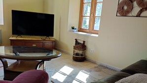Un salon avec un téléviseur à écran plat posé sur un meuble en bois, une table basse en verre, un panier en osier, un canapé et une fenêtre donnant sur un paysage verdoyant.