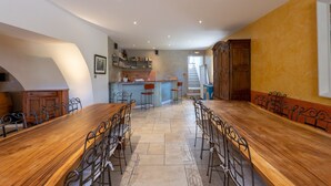 A spacious dining area with a long wooden table and metal chairs, a kitchen with a bar, and a staircase leading to an upper level. - Holiday Home “Bastide de Marius” with Shared Pool, Terrace, and Wi-Fi (Redortiers)