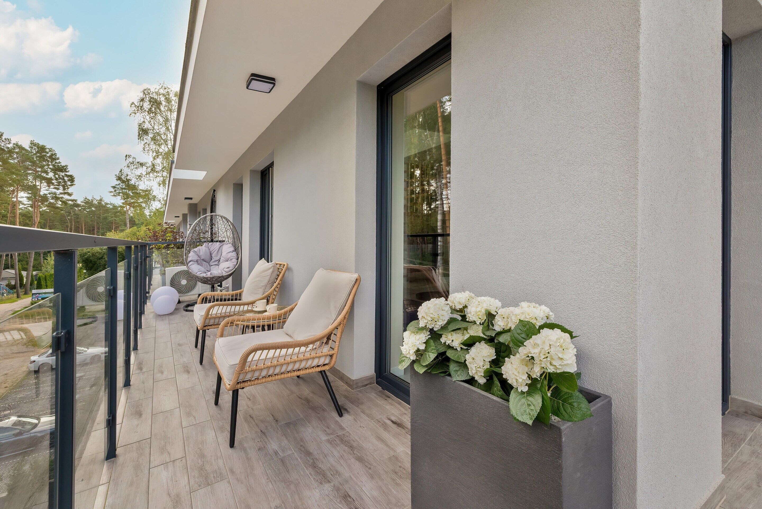 Un balcon avec des fauteuils en osier, une balustrade en verre et un jardinier orné de fleurs blanches.