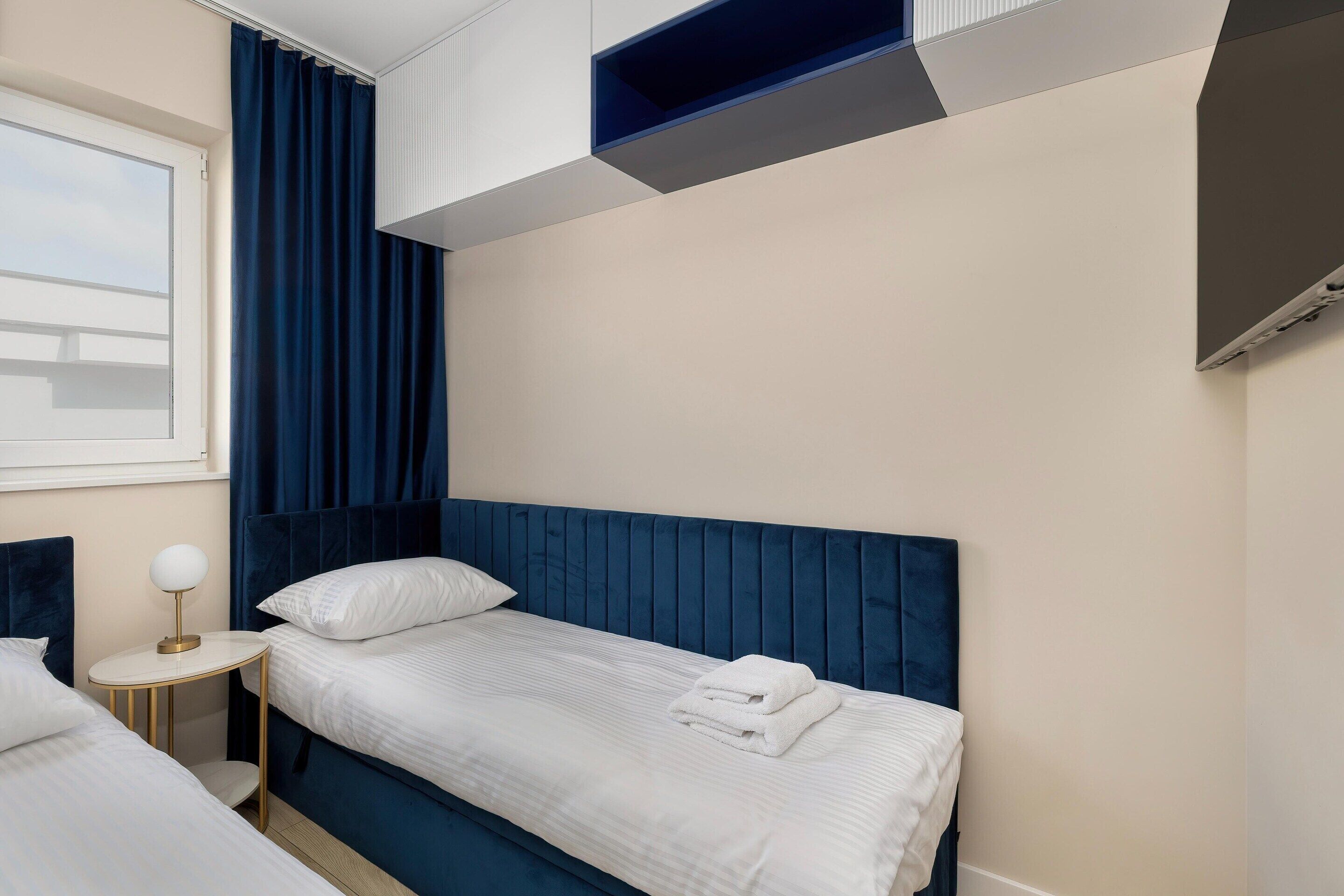 A hotel room with two beds, a small round table, a window with curtains, and a built-in shelf above the beds.