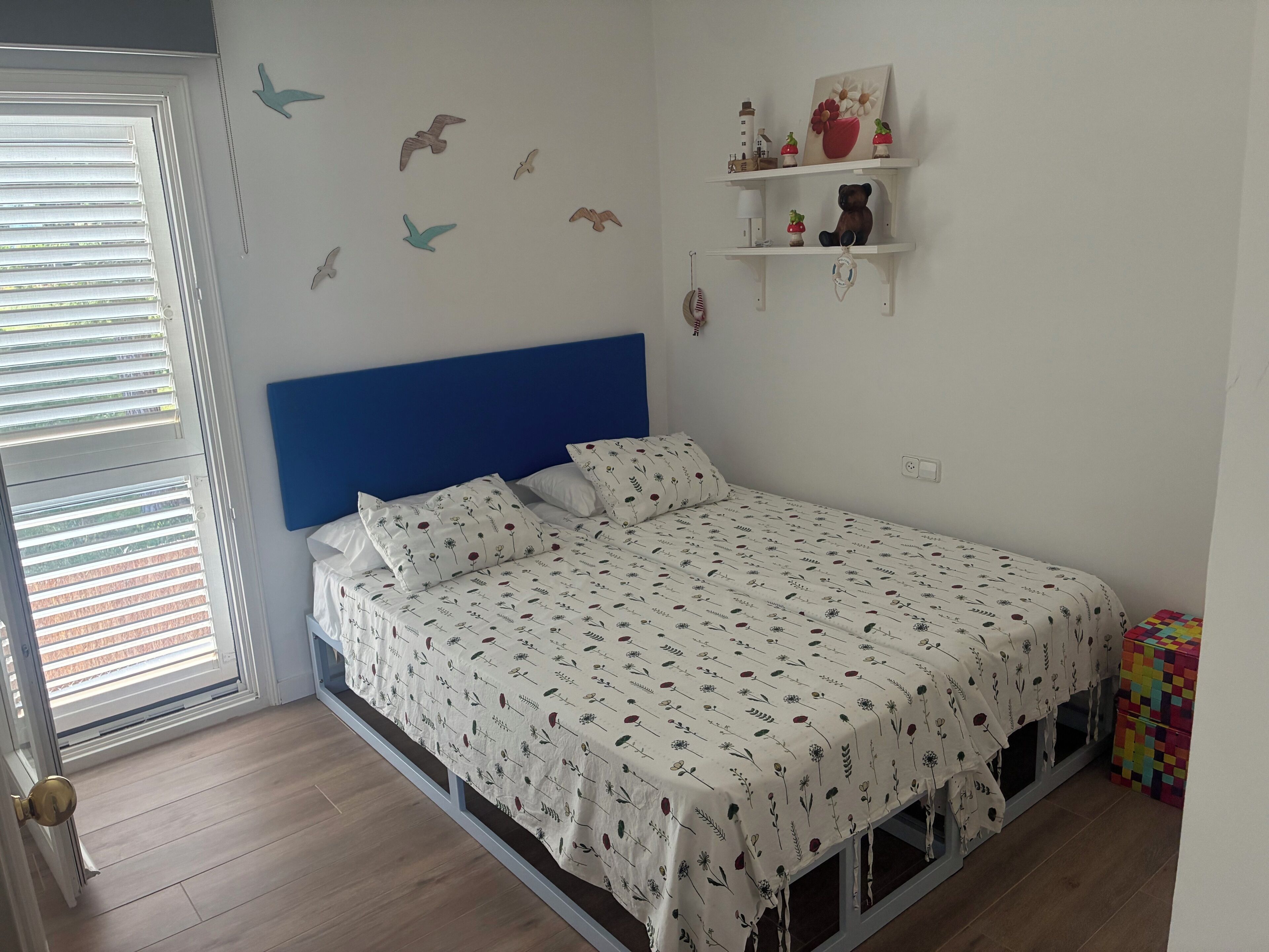 Une chambre à coucher avec un lit, une tête de lit bleue, une fenêtre avec des stores, un mur décoré d’autocollants représentant des oiseaux et une étagère ornée d’objets décoratifs.