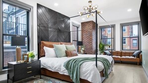 A modern bedroom with a large bed, a black headboard, a green throw blanket, and a view of the city through large windows. - North End 2-Story Penthouse / Prime Location (Boston)