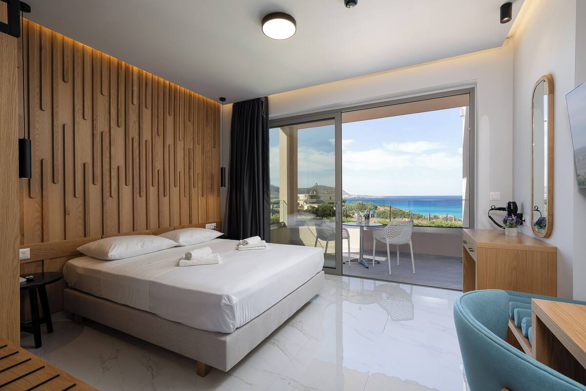 A modern hotel room with a large bed, a desk, a chair, a balcony with a view of the sea, and a wooden wall paneling.