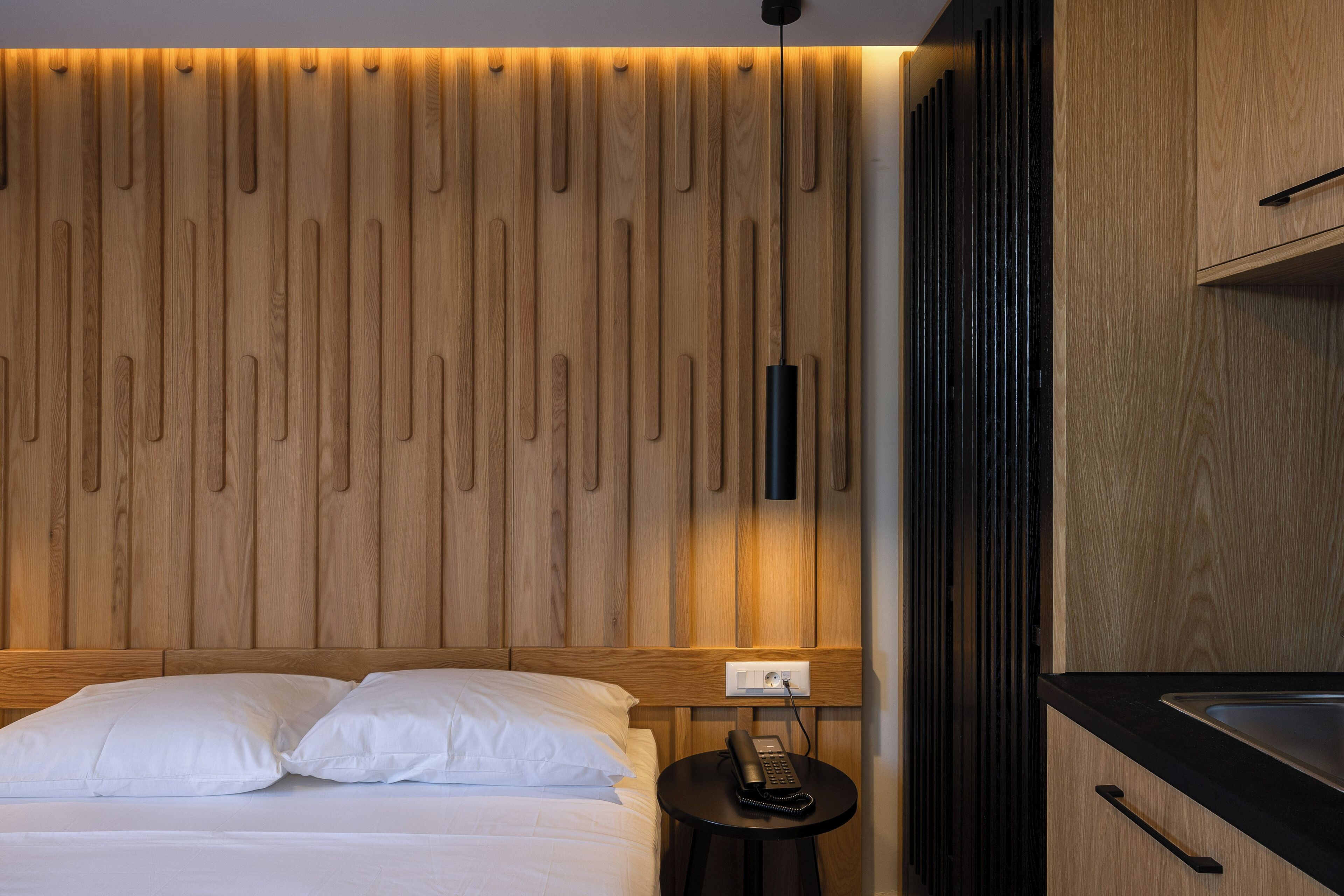 A hotel room with a wooden headboard, a bedside table with a phone, and a modern kitchen area.