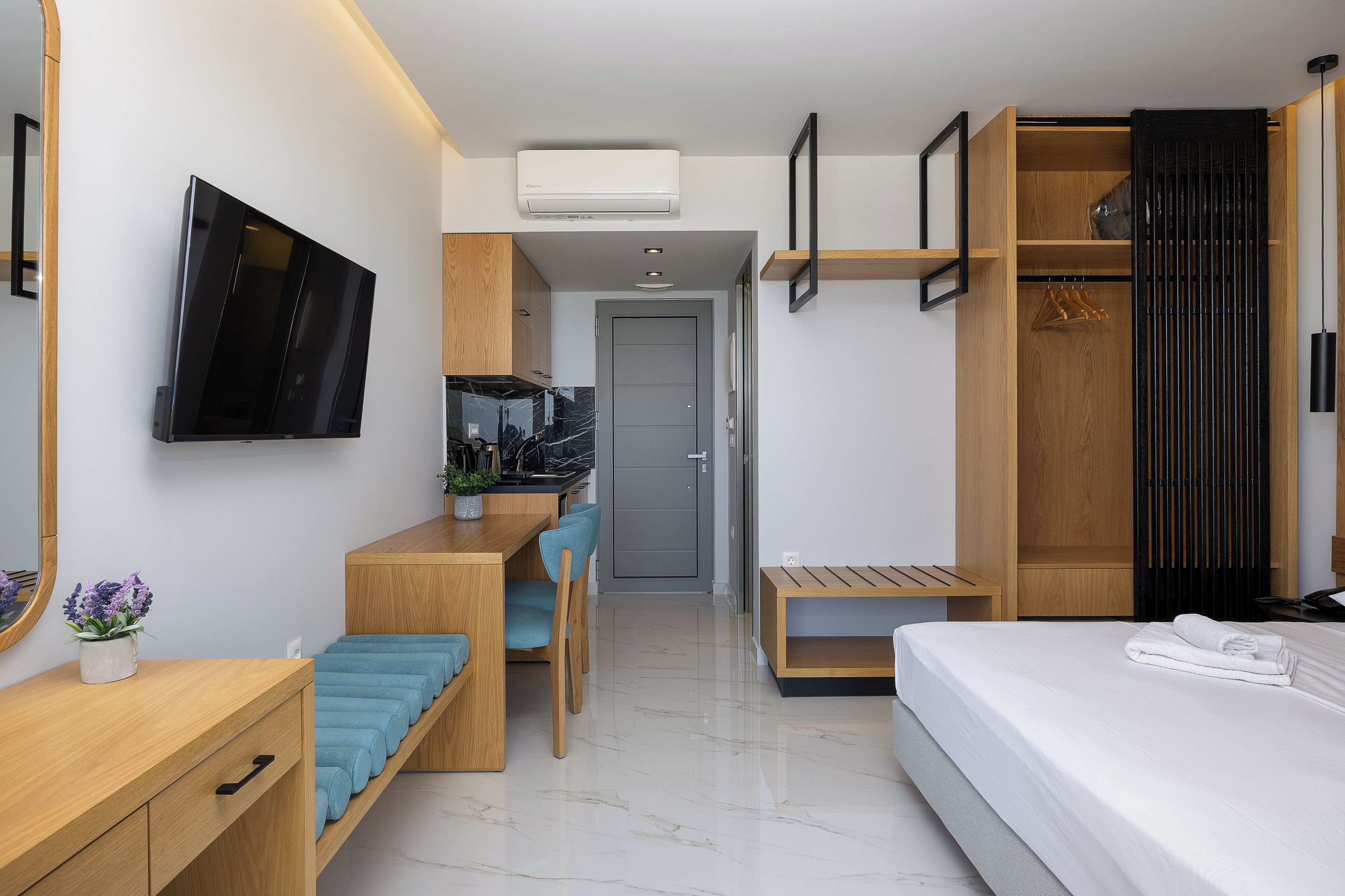 A modern hotel room with a flat-screen TV, a wooden desk with a chair, a bed with a neatly made white towel, and a wardrobe with a hanging rack.