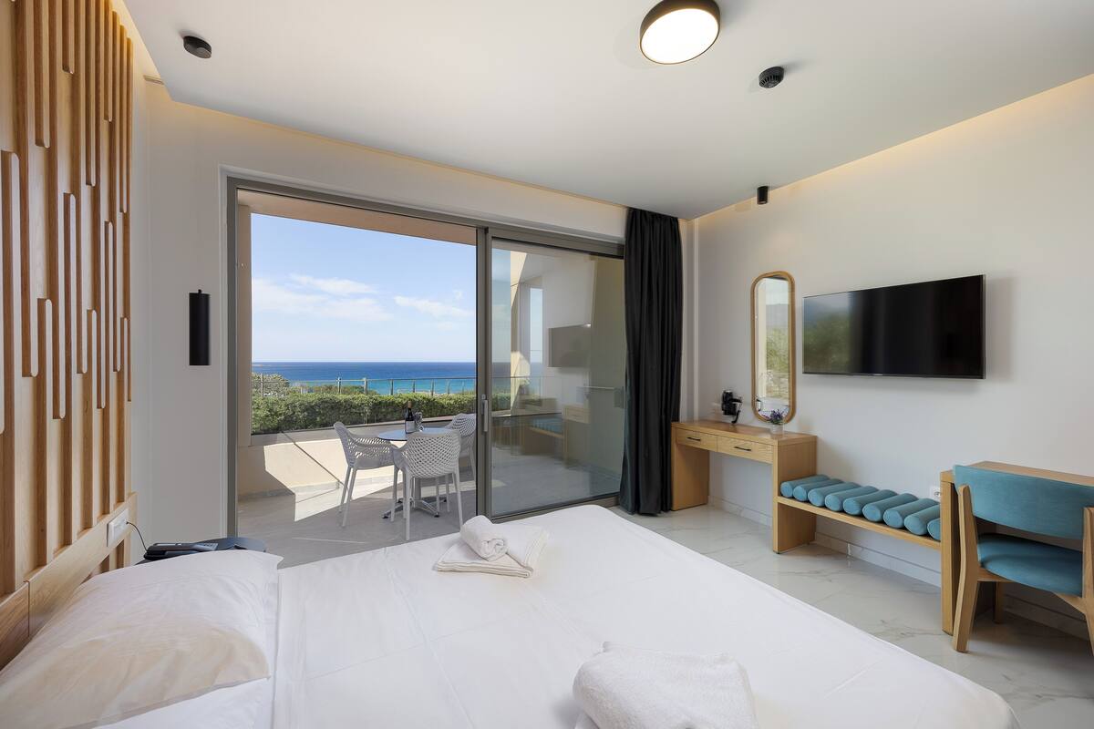 A hotel room with a bed, a desk, a TV, and a balcony with a view of the sea.