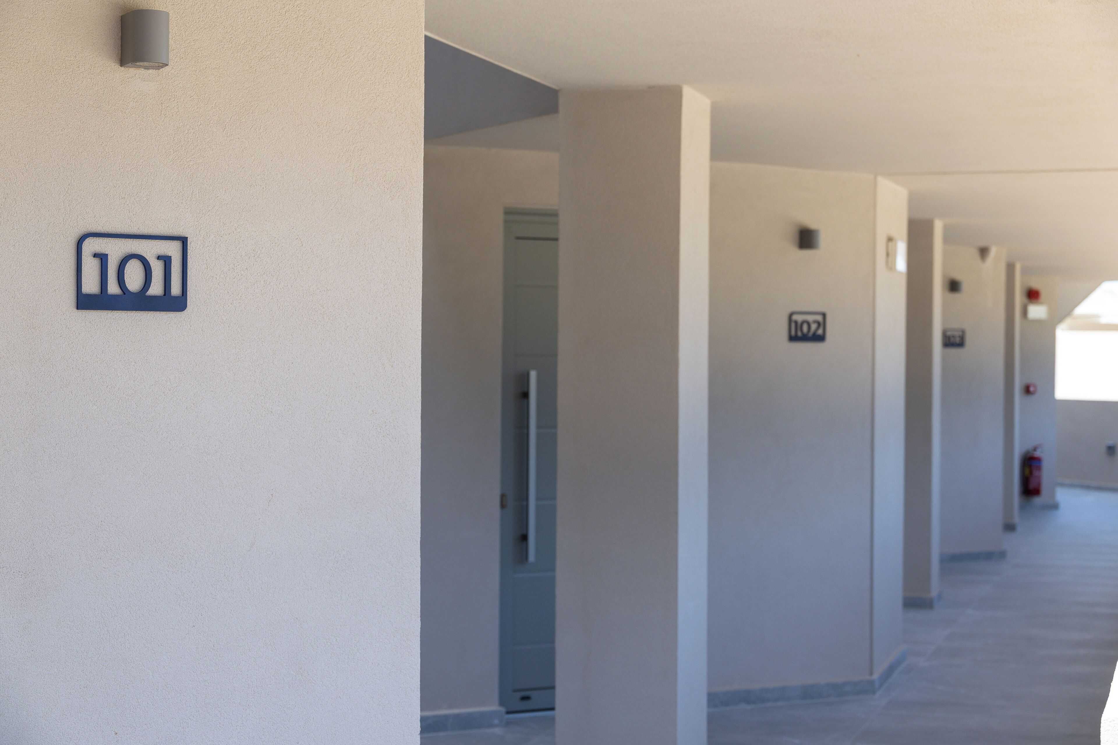 A corridor with numbered doors, a fire extinguisher, and a light fixture on the wall.