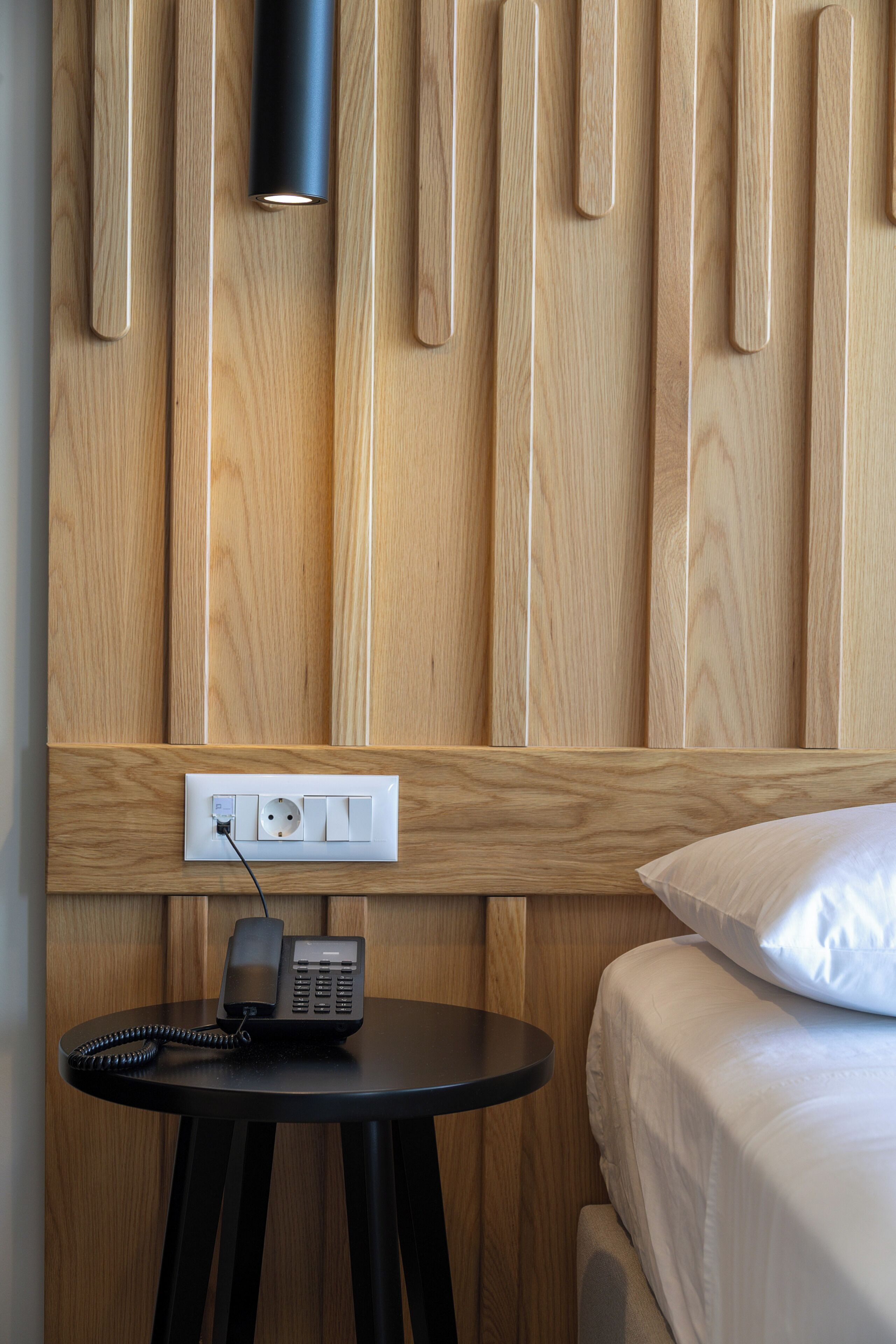 A hotel room with a wooden panel wall, a bedside table with a telephone, and a bed with white bedding.