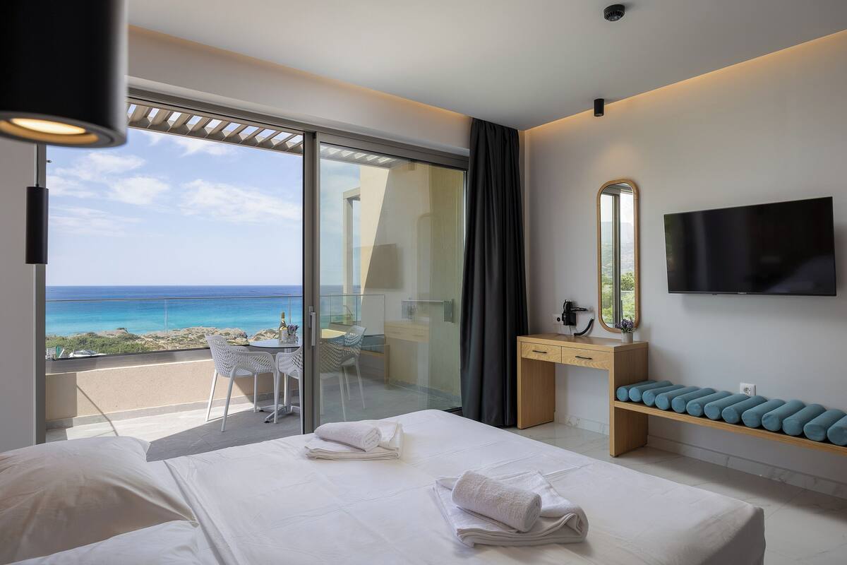 A hotel room with a bed, a desk, a chair, a TV, and a balcony with a view of the ocean.