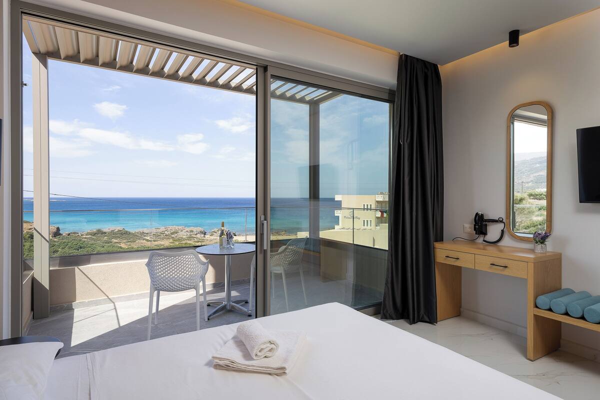 A hotel room with a bed, a desk, a chair, a small table, and a view of the ocean.