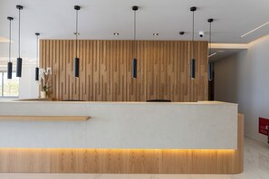 A reception desk with a wooden panel backdrop, pendant lighting, and a built-in bench. - Falasarna Bay Hotel (Kissamos)
